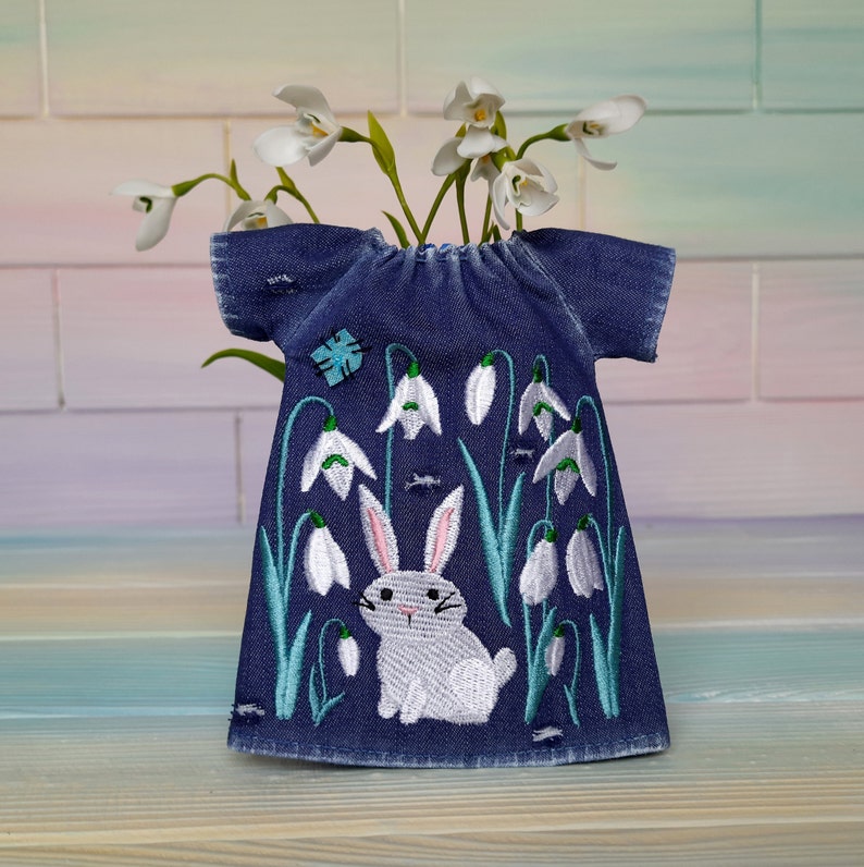 Puede incluir: Un vestido de mezclilla azul con campanillas blancas bordadas y un conejito blanco. El vestido tiene mangas cortas y un escote redondo.