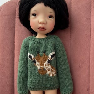 May include: A doll with black hair wearing a green sweater with a giraffe design. The doll has a fair complexion and is seated on a pink surface. The sweater has long sleeves and a detailed giraffe graphic.