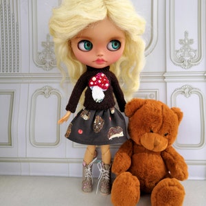 Puede incluir: Una mu&ntilde;eca con cabello rubio que lleva un su&eacute;ter marr&oacute;n con una aplicaci&oacute;n de hongo rojo y una falda negra y marr&oacute;n con un patr&oacute;n de erizo. La mu&ntilde;eca sostiene un oso de peluche marr&oacute;n.