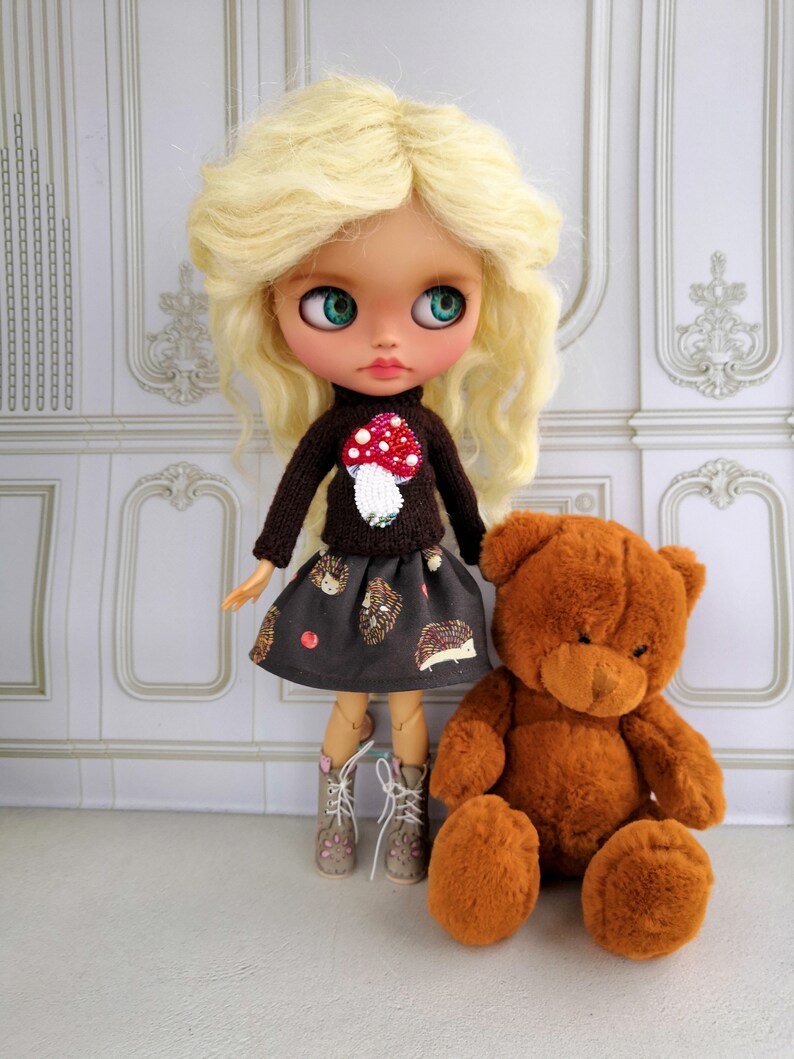 Puede incluir: Una mu&ntilde;eca con cabello rubio que lleva un su&eacute;ter marr&oacute;n con una aplicaci&oacute;n de hongo rojo y una falda negra y marr&oacute;n con un patr&oacute;n de erizo. La mu&ntilde;eca sostiene un oso de peluche marr&oacute;n.