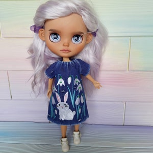 Puede incluir: Una mu&ntilde;eca con el pelo largo, ondulado y de color morado claro que lleva un vestido de mezclilla azul con flores blancas bordadas y un conejo blanco. La mu&ntilde;eca tiene ojos azules y una tez marr&oacute;n claro.