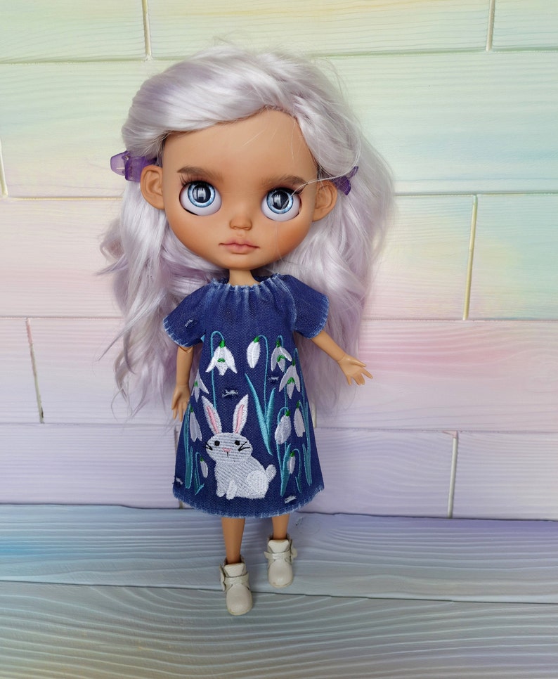 Puede incluir: Una mu&ntilde;eca con el pelo largo, ondulado y de color morado claro que lleva un vestido de mezclilla azul con flores blancas bordadas y un conejo blanco. La mu&ntilde;eca tiene ojos azules y una tez marr&oacute;n claro.