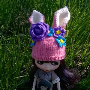 May include: A pink knitted hat with bunny ears and flower embellishments. The hat has purple, blue, and yellow flowers.