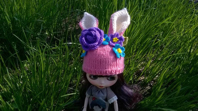 May include: A pink knitted hat with bunny ears and flower embellishments. The hat has purple, blue, and yellow flowers.