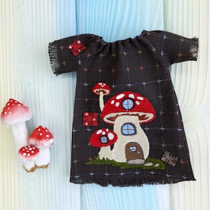 Può includere: Un vestito marrone con un designo di casa di funghi ricamato sul davanti. Il vestito ha un collo arricciato e un bordo a frangia. La casa di funghi ha un cappello rosso con macchie bianche e un gambo marrone. Ci sono due funghi più piccoli su entrambi i lati della casa di funghi principale.