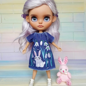 Puede incluir: Una mu&ntilde;eca con el pelo largo, ondulado y lavanda que lleva un vestido de mezclilla azul con flores blancas bordadas y un conejito. La mu&ntilde;eca sostiene un conejito de peluche rosa con un lazo blanco.