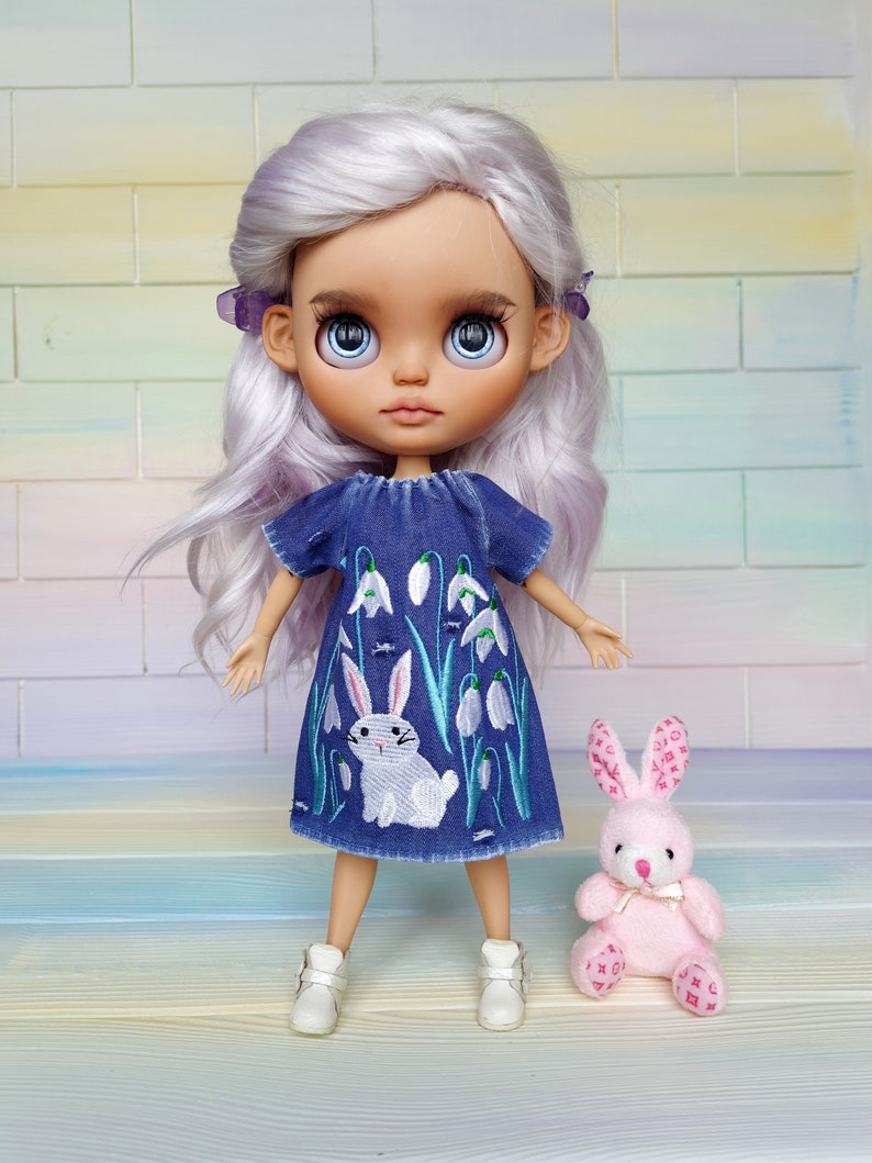 Puede incluir: Una mu&ntilde;eca con el pelo largo, ondulado y lavanda que lleva un vestido de mezclilla azul con flores blancas bordadas y un conejito. La mu&ntilde;eca sostiene un conejito de peluche rosa con un lazo blanco.