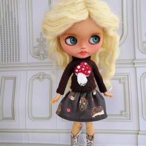 Puede incluir: Una mu&ntilde;eca con cabello rubio que lleva un su&eacute;ter marr&oacute;n con un dise&ntilde;o de hongo y una falda marr&oacute;n con un patr&oacute;n de erizo. La mu&ntilde;eca lleva botas marrones con detalles rosas.