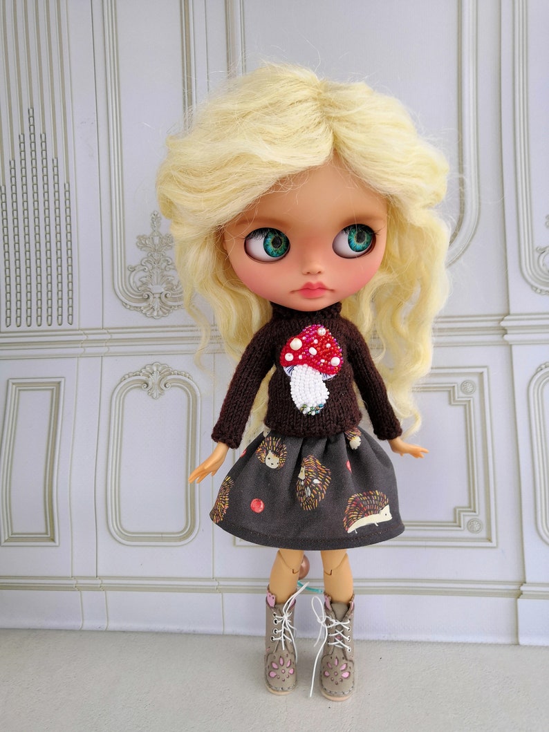 Puede incluir: Una mu&ntilde;eca con cabello rubio que lleva un su&eacute;ter marr&oacute;n con un dise&ntilde;o de hongo y una falda marr&oacute;n con un patr&oacute;n de erizo. La mu&ntilde;eca lleva botas marrones con detalles rosas.