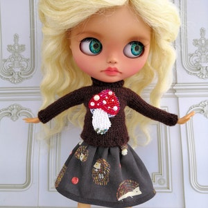 Puede incluir: Una mu&ntilde;eca con cabello rubio que lleva un su&eacute;ter marr&oacute;n con un dise&ntilde;o de hongo rojo y blanco y una falda marr&oacute;n con un patr&oacute;n de erizo.