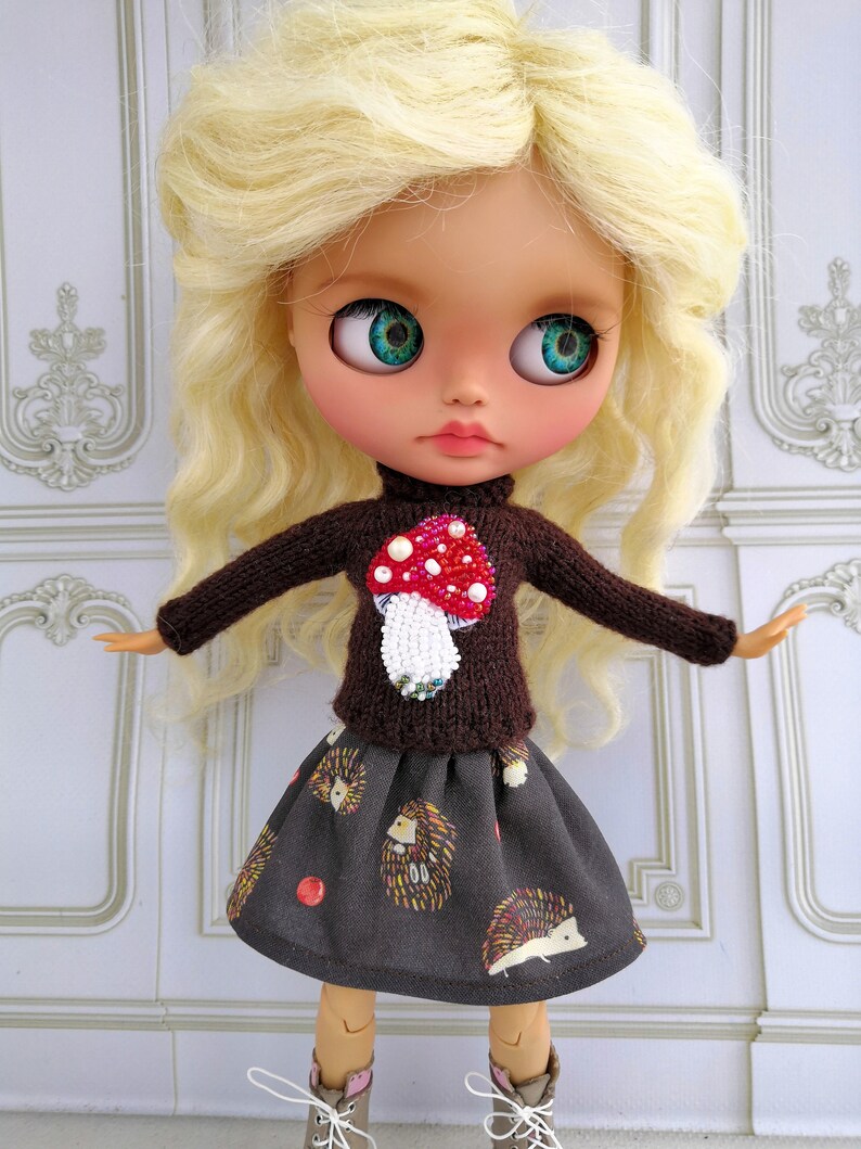 Puede incluir: Una mu&ntilde;eca con cabello rubio que lleva un su&eacute;ter marr&oacute;n con un dise&ntilde;o de hongo rojo y blanco y una falda marr&oacute;n con un patr&oacute;n de erizo.