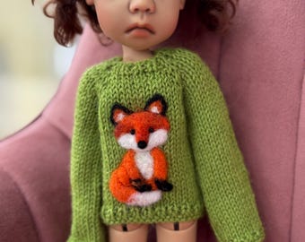 Op de afbeelding: Een pop met bruin haar in staartjes draagt een groen gebreide trui met lange mouwen. De trui is voorzien van een naaldgevilte afbeelding van een vos in de kleuren oranje, wit en zwart op het voorpand.