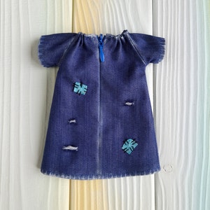 Puede incluir: Un vestido de mezclilla azul con cierre de cremallera y dos parches azules y turquesa. El vestido tiene un aspecto desgastado con varios agujeros peque&ntilde;os.