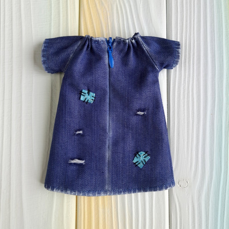 Puede incluir: Un vestido de mezclilla azul con cierre de cremallera y dos parches azules y turquesa. El vestido tiene un aspecto desgastado con varios agujeros peque&ntilde;os.