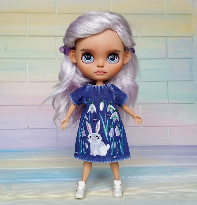 Puede incluir: Una mu&ntilde;eca con el pelo largo y blanco que lleva un vestido de mezclilla azul con flores blancas bordadas y un conejito blanco. La mu&ntilde;eca tiene ojos azules y lleva zapatos blancos.