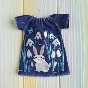 Puede incluir: Un vestido de mezclilla azul con campanillas blancas bordadas y un conejito blanco. El vestido tiene un escote fruncido y mangas cortas.