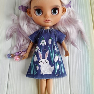 Puede incluir: Una mu&ntilde;eca con el pelo largo, ondulado y lavanda que lleva un vestido de mezclilla azul con un conejo blanco y flores blancas bordadas. La mu&ntilde;eca lleva zapatos blancos.