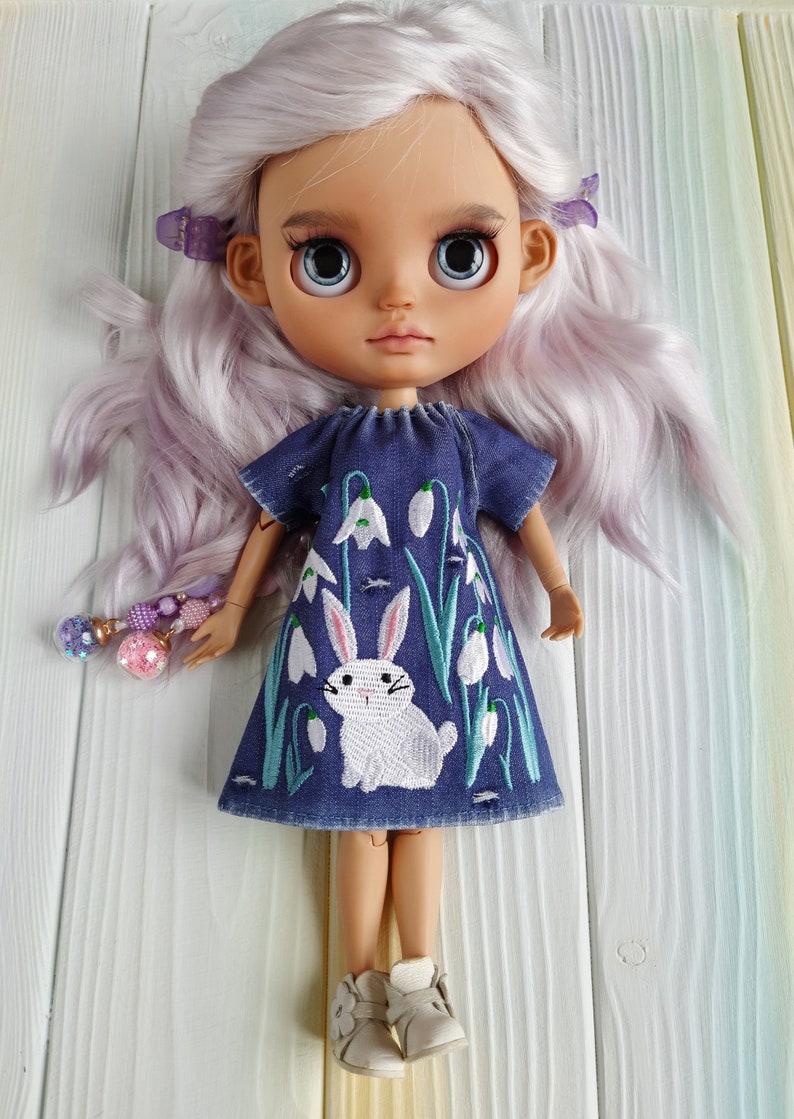 Puede incluir: Una mu&ntilde;eca con el pelo largo, ondulado y lavanda que lleva un vestido de mezclilla azul con un conejo blanco y flores blancas bordadas. La mu&ntilde;eca lleva zapatos blancos.