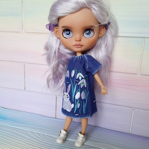Puede incluir: Una mu&ntilde;eca con el pelo largo y blanco que lleva un vestido de mezclilla azul con flores blancas bordadas y un conejito blanco. La mu&ntilde;eca tiene ojos azules y lleva zapatos blancos.
