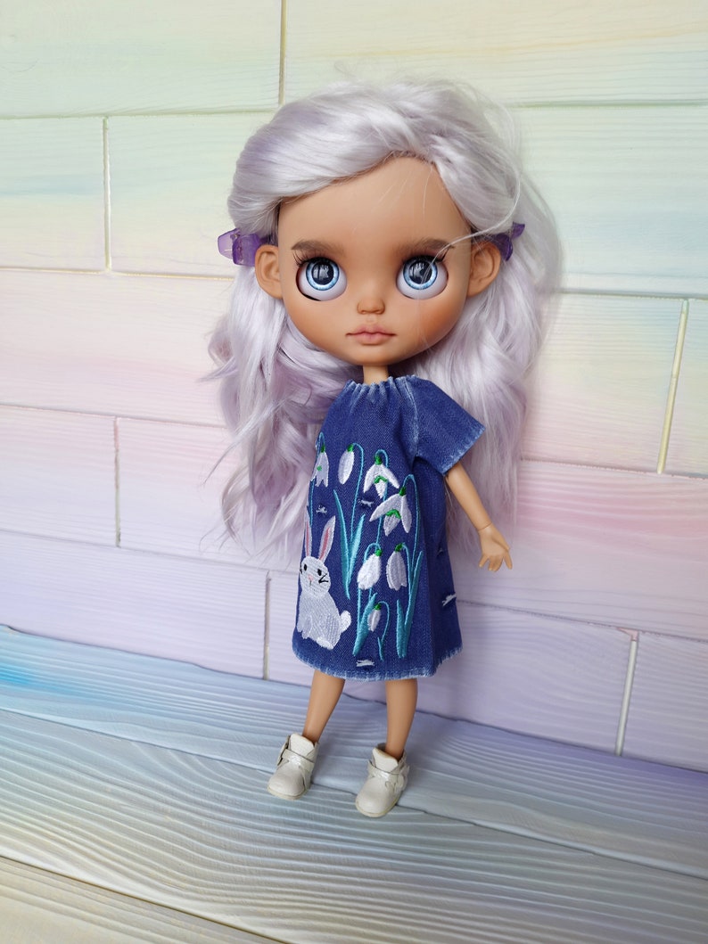 Puede incluir: Una mu&ntilde;eca con el pelo largo y blanco que lleva un vestido de mezclilla azul con flores blancas bordadas y un conejito blanco. La mu&ntilde;eca tiene ojos azules y lleva zapatos blancos.