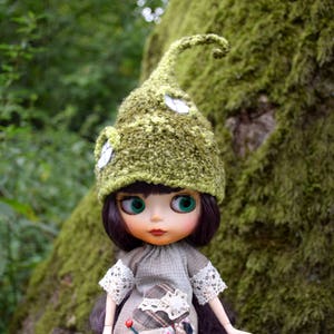 Puede incluir: Una muñeca con un gorro verde tejido con detalles blancos y un vestido gris con mangas de encaje. La muñeca tiene cabello oscuro, ojos verdes grandes y está frente a un tronco de árbol cubierto de musgo.