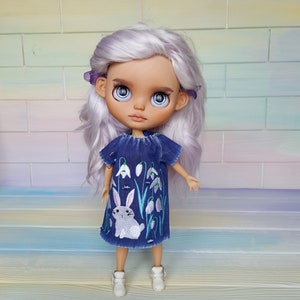 Puede incluir: Una mu&ntilde;eca con el pelo largo y blanco que lleva un vestido de mezclilla azul con flores blancas bordadas y un conejo blanco. La mu&ntilde;eca lleva zapatos blancos y tiene ojos azules.