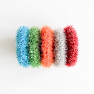 May include: Four colorful scrubbers made of a soft, fluffy material. The scrubbers are arranged in a row from left to right: blue, green, orange, and red.