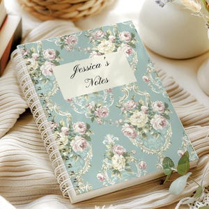 Könnte beinhalten: Ein Notizbuch mit floralem Design und Spiralbindung. Das Cover ist hellblau mit rosa und weißen Rosen und goldenen Akzenten. Der Text "Jessica's Notes" befindet sich auf einem cremefarbenen Etikett.