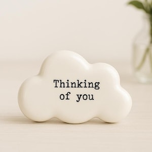 May include: A white cloud-shaped ceramic ornament with the text "Thinking of you" in black. The ornament is on a light-coloured surface, with a small glass vase of flowers in the background.
