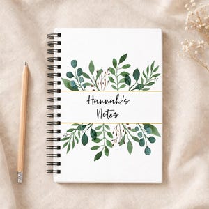 Op de afbeelding: Een wit spiraalgebonden notitieboek met de tekst "Hannah's Notes", versierd met groene aquarelbladeren en gouden lijnen. Een houten potlood ligt naast het notitieboek. Het notitieboek is ongeveer 21 cm lang.
