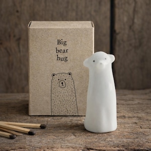 May include: A white ceramic bear figurine with a rounded body and small ears. The figurine is next to a cardboard box with the text "Big bear hug" and a bear illustration. Matches are in the foreground.