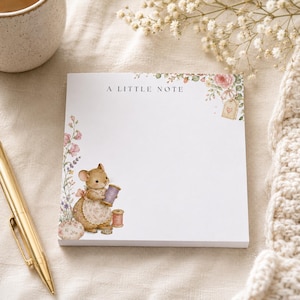 May include: A square notepad with the text A LITTLE NOTE printed at the top. The paper features a watercolour illustration of a mouse wearing an apron, holding a purple spool of thread, surrounded by floral accents and a small gift tag.