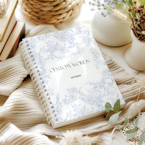 May include: A white spiral-bound notebook with a blue floral pattern and the words "CUSTOM WORDS" and "NOTEBOOK". The notebook is open, revealing blank pages. The background includes beige fabric and decorative items.