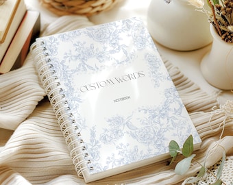 Puede incluir: Un cuaderno de espiral blanco con una cubierta con diseño floral azul y las palabras "CUSTOM WORDS" y "NOTEBOOK". El cuaderno está abierto, mostrando páginas en blanco. El fondo incluye tela beige y elementos decorativos.