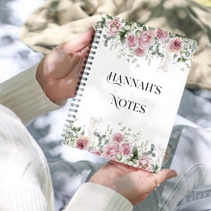 May include: A spiral-bound notebook with the text "HANNAH'S NOTES" in elegant script. The cover features a floral design with pink roses and green foliage. The notebook is held in a person's hands.