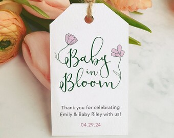 May include: A white gift tag with the words "Baby in Bloom" in green script, adorned with pink flower illustrations. The tag includes the text "Thank you for celebrating Emily & Baby Riley with us!" and the date 04.29.24.