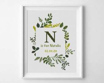 May include: Framed cross-stitch artwork featuring a green letter "N" inside a square, surrounded by a wreath of green leaves. The text "is for Natalie" and the date "02.08.20" are below the letter. The artwork is in a white frame.