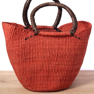 Op de afbeelding: Een grote, rode, geweven mand met twee bruine leren handvatten. De mand is gemaakt van een natuurlijke vezel en heeft een licht afgeronde vorm.