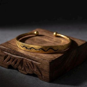 Peut inclure: Bracelet manchette doré avec un motif de vagues noires. Le bracelet ouvert présente de petits accents de boules aux extrémités. Le bracelet est présenté sur un bloc de bois sculpté, mettant en valeur son design et son savoir-faire.