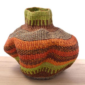 Puede incluir: Una cesta tejida con forma redonda y bulbosa. La cesta está hecha de fibras naturales y tiene un patrón de rayas coloridas en tonos marrones, naranjas y verdes.