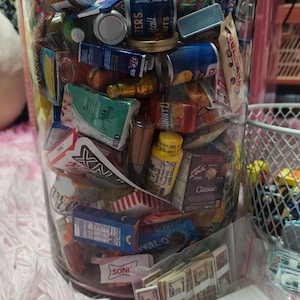 May include: A clear cylindrical container filled with a variety of miniature toy food items, including small cans, boxes, and bottles with brand labels like Skippy, SunnyD, and Whoppers, alongside small plastic bags containing miniature toy currency.