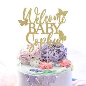 May include: Gold glitter cake topper with the text "Welcome Baby Sophia" and butterflies. The topper is on a purple and pink frosted cake.