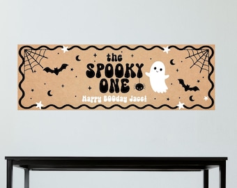May include: A rectangular tan banner featuring black wavy borders, spiderwebs, bats, stars, and moons. The text reads "the SPOOKY ONE" in a bold, retro font, with a small white ghost and a spider hanging from the text, plus "Happy 800day Jace!"