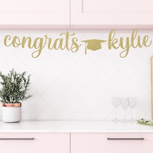 May include: Gold glitter graduation banner with the text "congrats kylie" and a graduation cap.