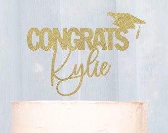 May include: Gold glitter cake topper with the text "Congrats Kylie" and a graduation cap. The topper is on top of a white frosted cake.