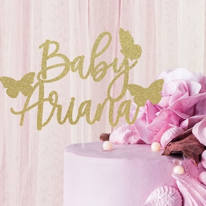 May include: A pink cake with a gold glitter topper that says "Baby Ariana" with butterfly accents. The cake is decorated with pink frosting and chocolate accents.