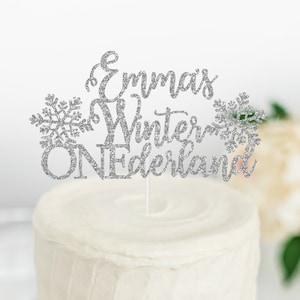 May include: Silver glitter cake topper with the text "Emma's Winter Wonderland One" and snowflake designs.