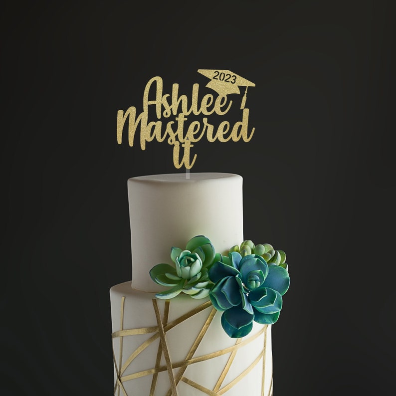 K&ouml;nnte beinhalten: Eine zweist&ouml;ckige wei&szlig;e Torte mit goldenen geometrischen Designs und Sukkulenten-Dekorationen. Ein goldener Glitzer-Kuchenaufsatz mit der Aufschrift "Ashlee Mastered it" mit einer Abschlusskappe und dem Jahr 2023.