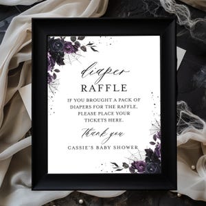 May include: A black picture frame containing a white sign with dark purple roses, spider webs, and black text that reads: diaper RAFFLE IF YOU BROUGHT A PACK OF DIAPERS FOR THE RAFFLE, PLEASE PLACE YOUR TICKETS HERE. Thank you CASSIE'S BABY SHOWER.