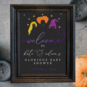 May include: A black and white welcome sign with a purple and gold glitter design featuring three witches. The sign reads "Welcome to Kate & Adam's Glorious Baby Shower October 4, 2022".